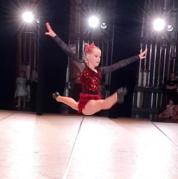 A young girl in a red sequined dress performing a jump on a dance stage with studio lights overhead.