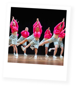 A group of young dancers on stage wearing pink tops and blue shorts, doing hip hop.