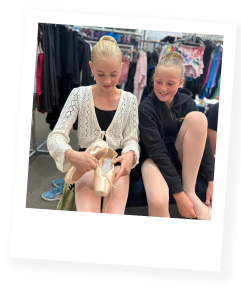 Two young girls sitting on a bench in a clothing store, one is trying on a pair of high heels while the other laughs.