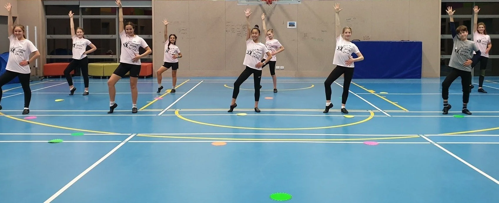 Group of young girls practicing dance in a gymnasium, standing in a line with hands on their hips, wearing matching t-shirts and black leggings.