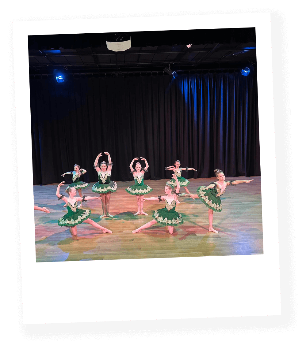 Young ballet dancers in green costumes performing a dance on stage with black curtains and stage lighting.