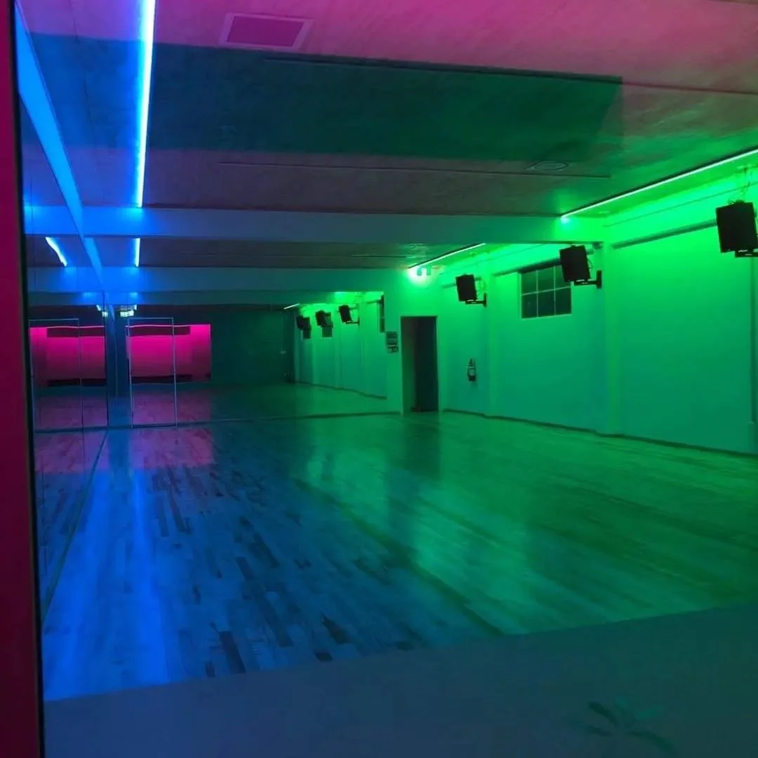 An empty dance studio with colorful LED lights illuminating the walls and ceiling, including blue, pink, and green. The room has a wooden floor and a large mirror on one side.