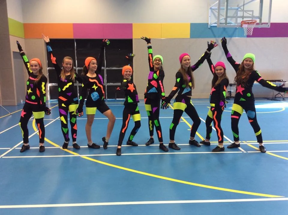 Group of nine young girls in colorful, glow-in-the-dark costumes, posing playfully with arms raised in a gymnasium.