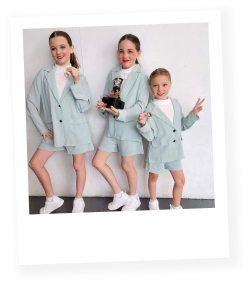 Three young girls dressed in light blue jackets, holding a trophy, and posing in front of a plain white background. They are smiling and making peace signs.