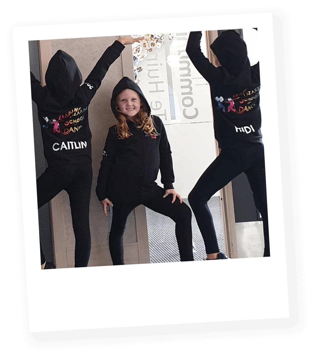 Three young girls in black hoodies and black pants posing in front of a wall, one girl smiling in the center and the other two girls on either side with their hoods up, one with their hand raised, all in a dance studio.