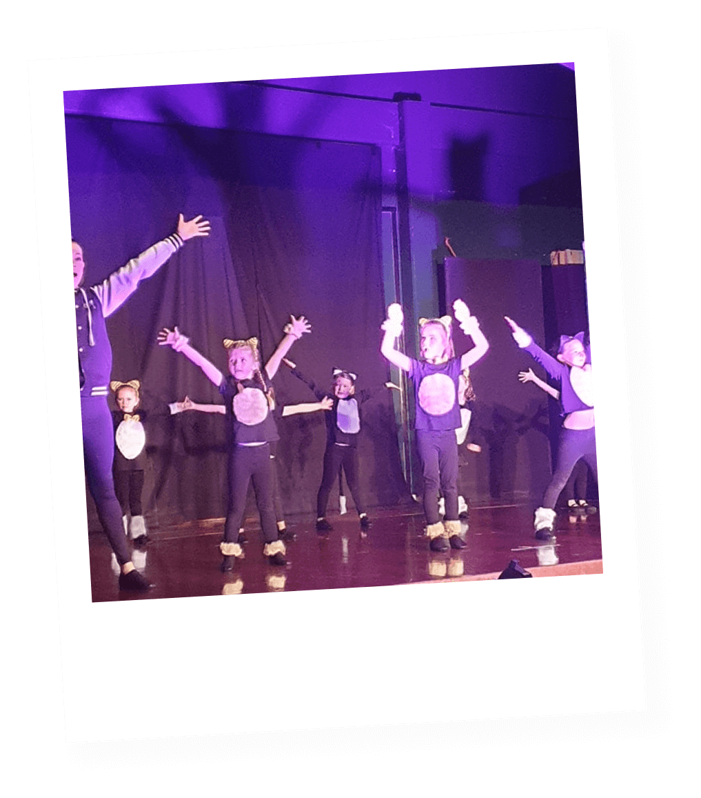 Children dressed as cats performing on stage with a dark backdrop, under purple stage lighting.