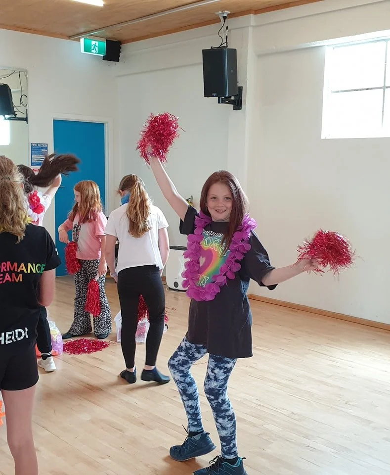 Young girl dancing and smiling at dance practice, holding red pom-poms, wearing a purple lei, black t-shirt, and blue patterned leggings.