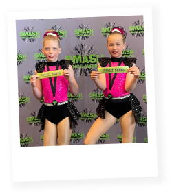 Two girls wearing pink and black dance costumes holding silver award ribbons and posing for the camera