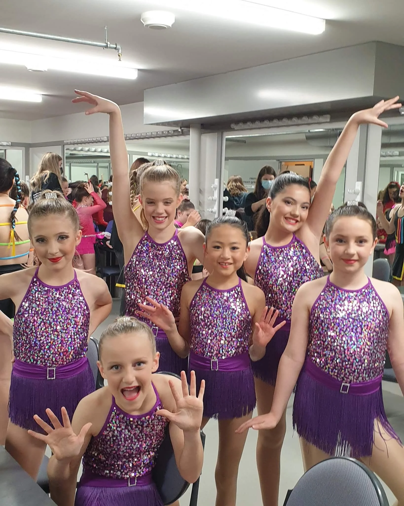 Group of young female dancers in purple sequined costumes posing backstage at a dance competition.