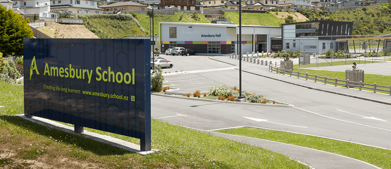 Signboard for Amesbury School with the slogan 'Creating life-long learners' and the school's website URL, near school buildings and parking lot.