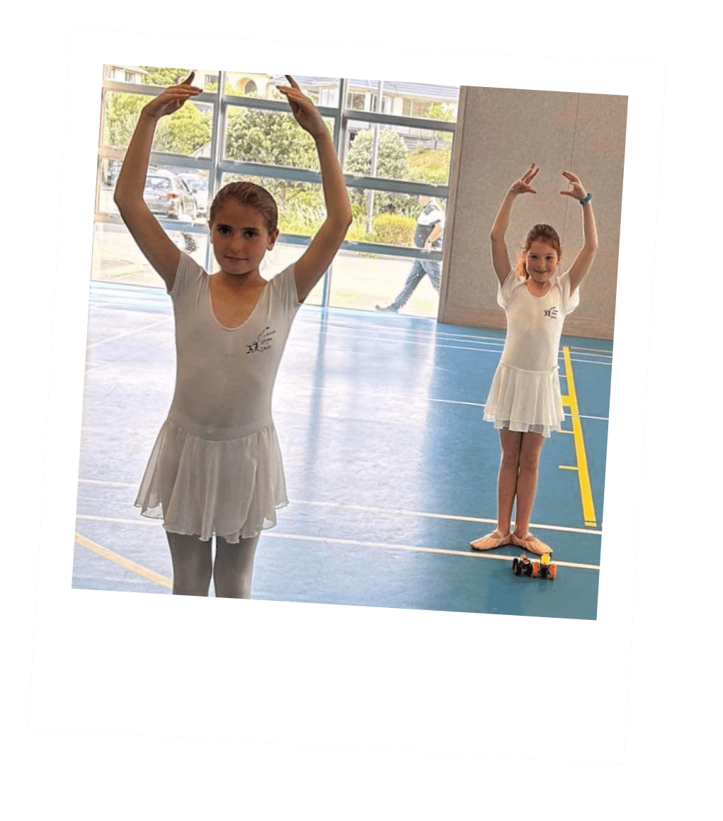 Two young girls in white ballet dresses standing on a gymnasium floor with arms raised, smiling, with large windows showing outside views.