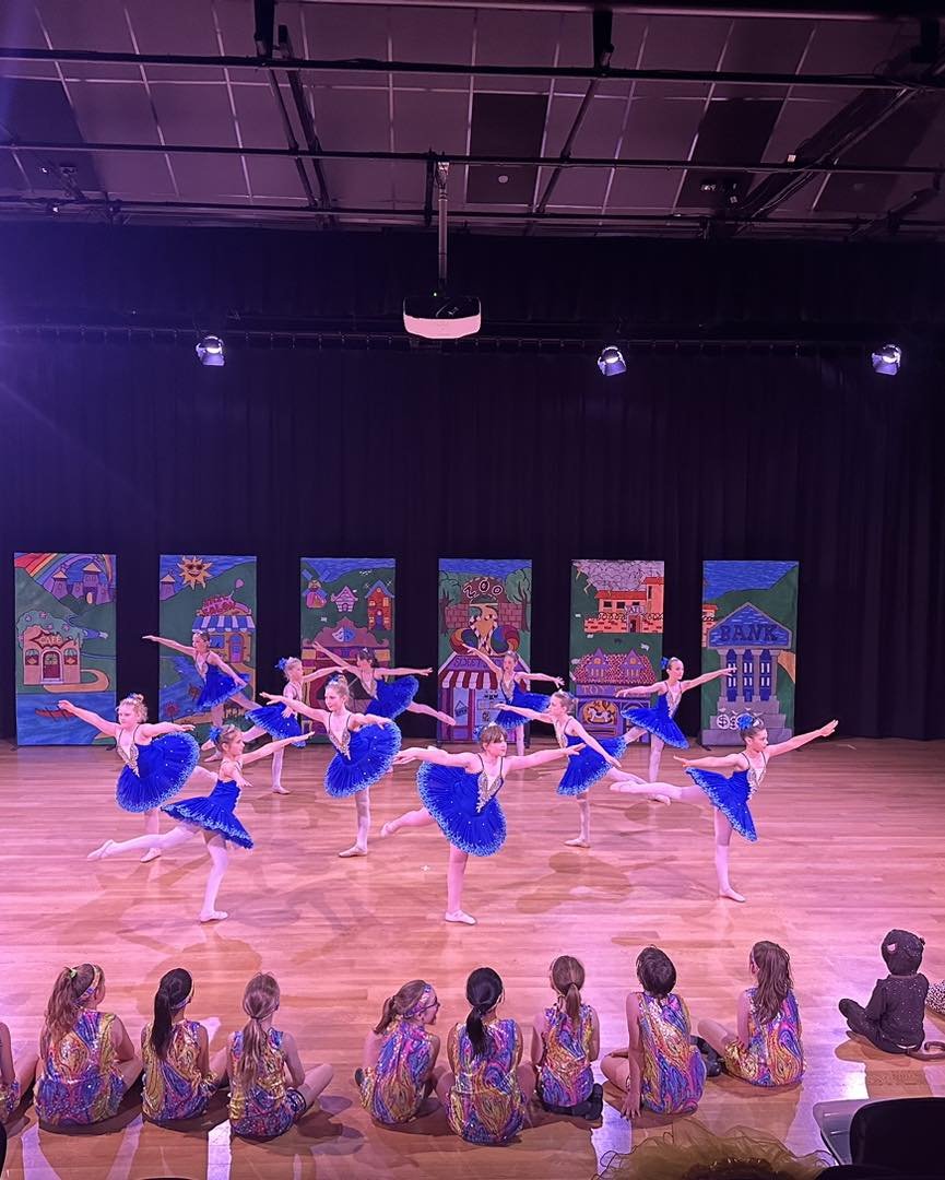 Young ballet dancers in blue dresses perform on stage during a recital, with an audience of children seated on the floor watching.