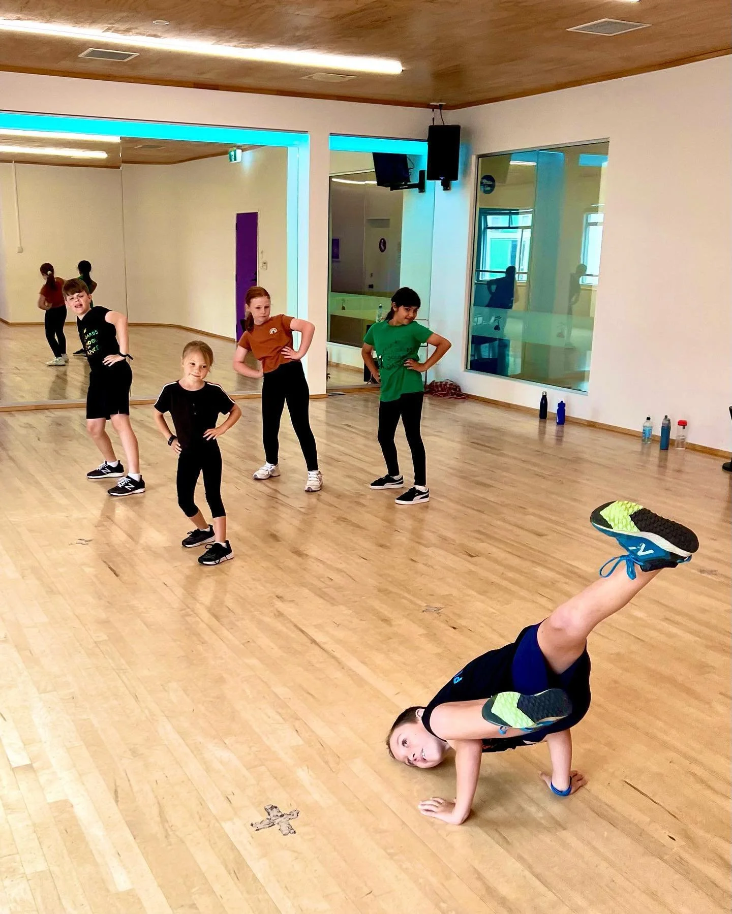 Group of children in a dance studio, with a boy doing a handstand in the foreground, and four children standing with hands on hips in the background.