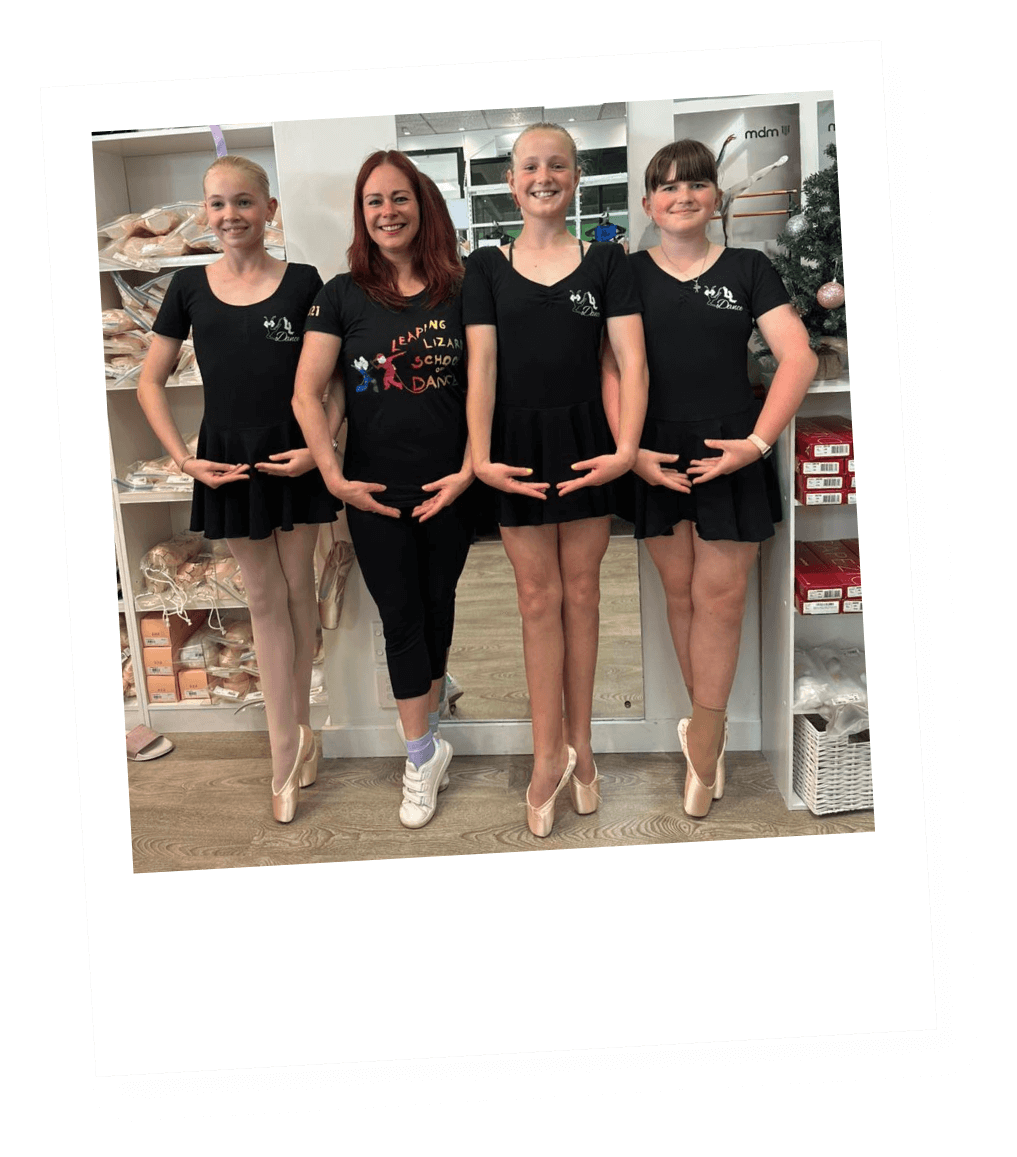 Four young girls and an adult woman standing in a dance studio, all wearing black dance costumes and ballet pointe shoes, posing with their hands in a ballerina pose.