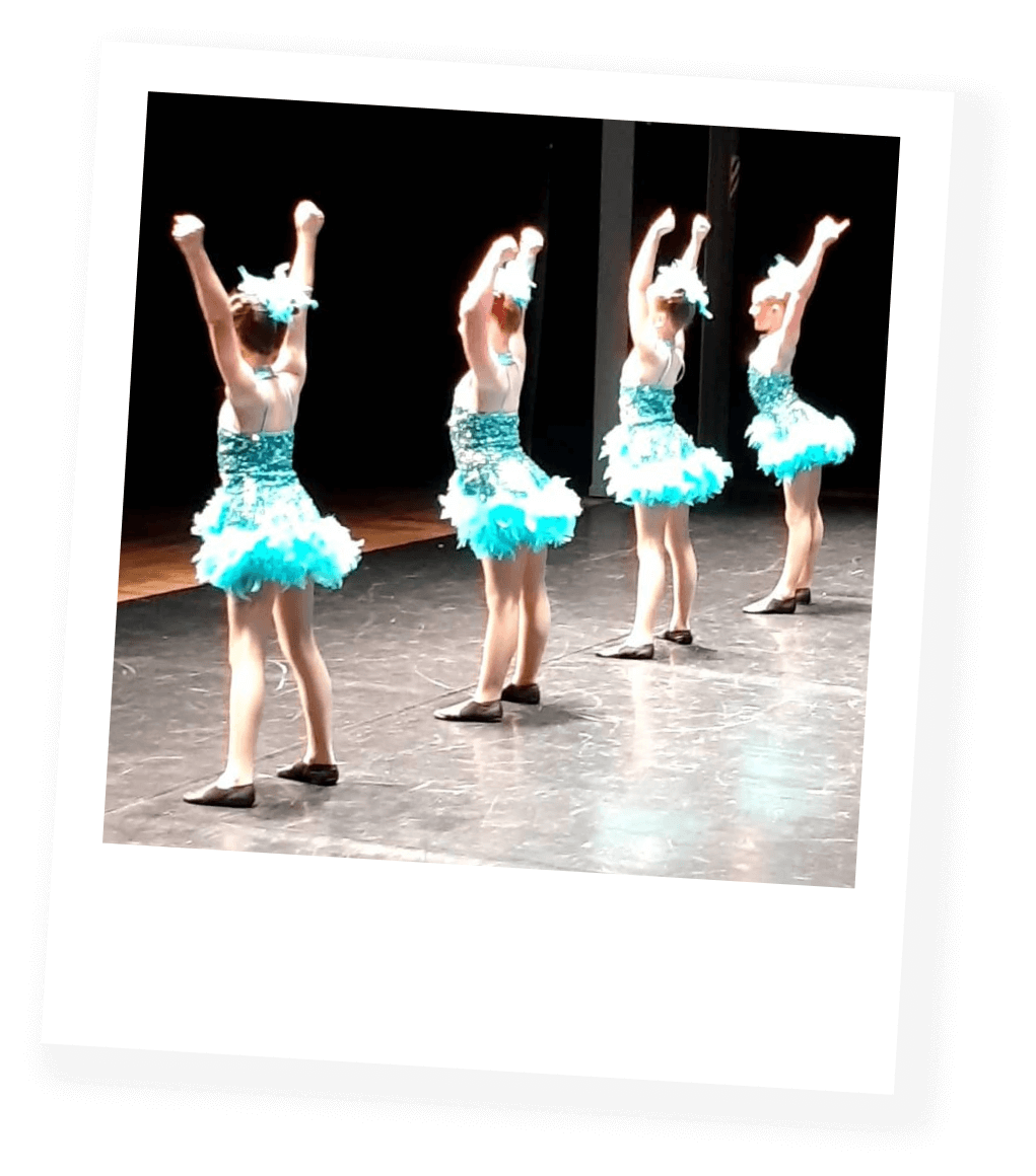 Four young girls in matching blue tutu dance costumes perform a choreographed dance on stage with their arms raised.