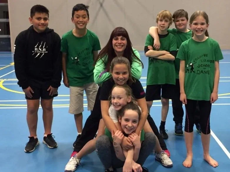 Group of children and a young woman smiling in a gymnasium, some wearing green T-shirts with 'Leaping Lizards School of Dance' logo.