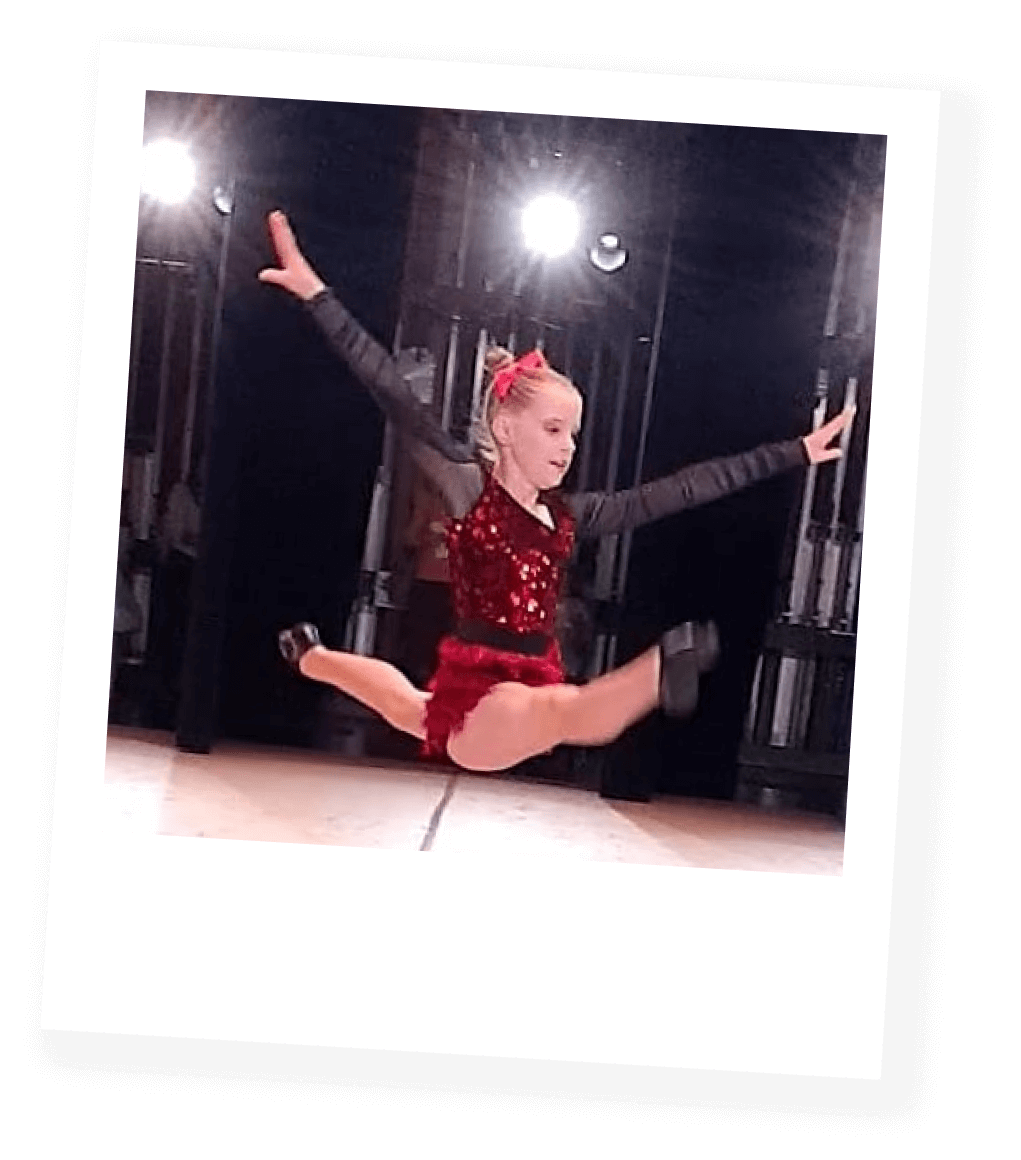 Young girl performing a split jump on stage with arms outstretched, wearing a red and black dance costume and hair accessory.
