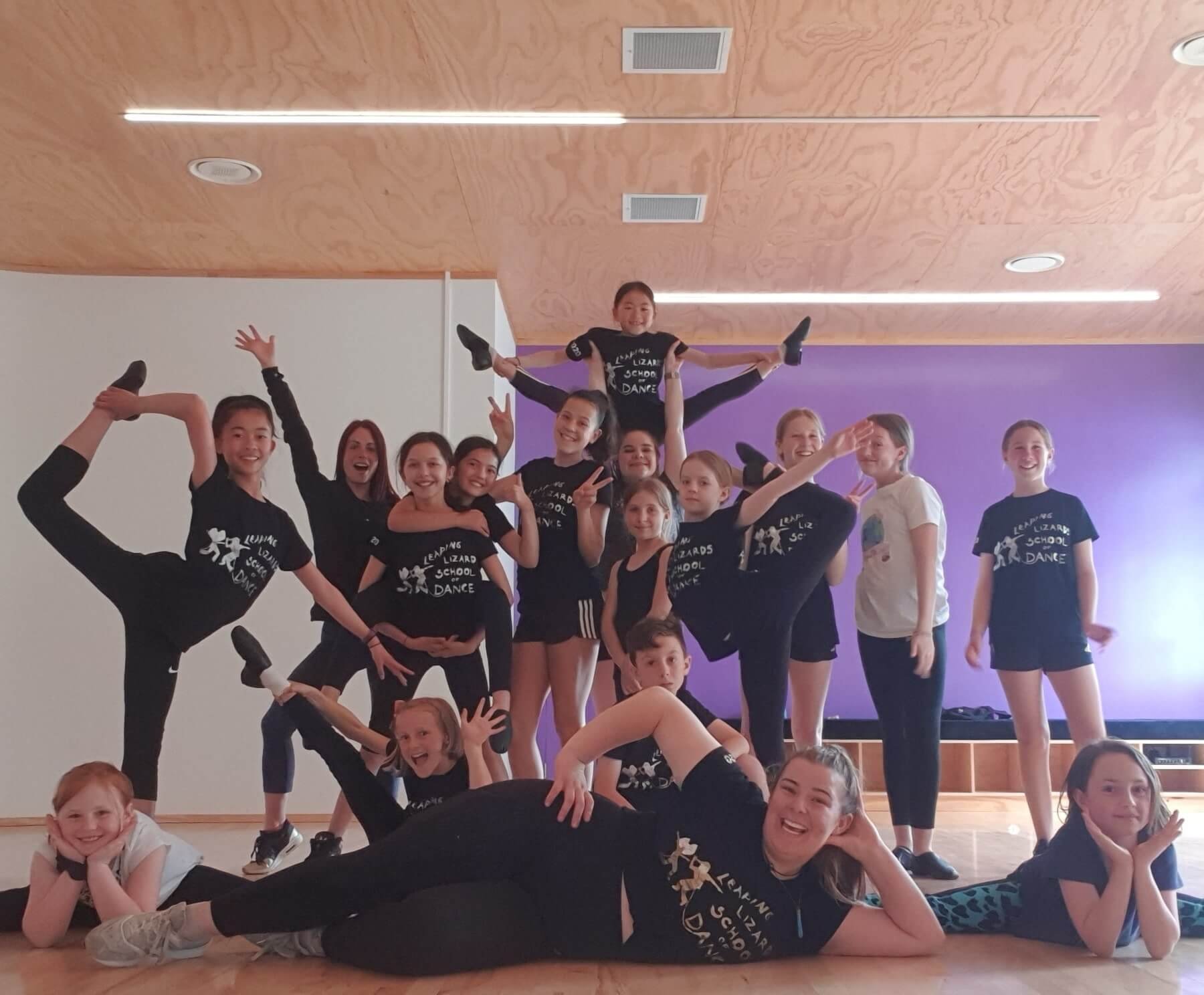 Group of children and teenagers, some in balancing poses, posing for a photo in a dance studio, with a purple wall and wooden ceiling.