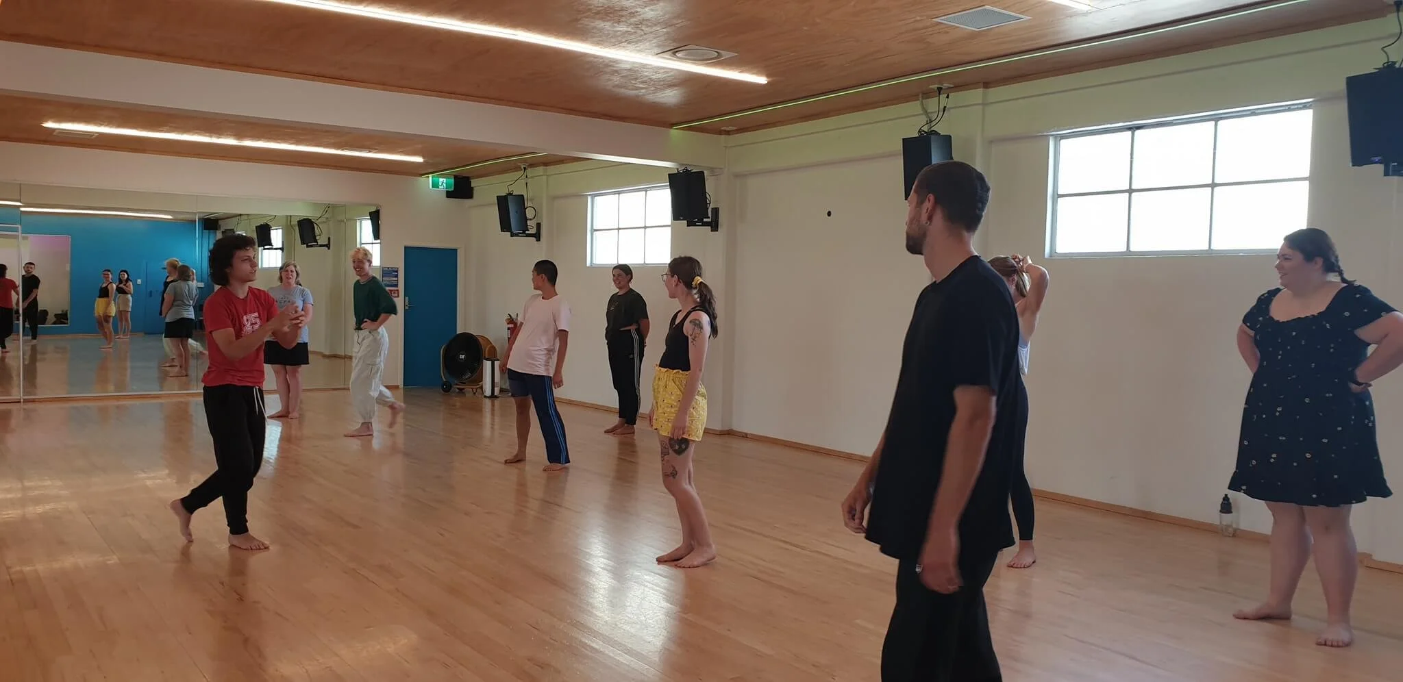 Group of people practicing dance or exercise in a studio with wood flooring, large mirror, and windows.