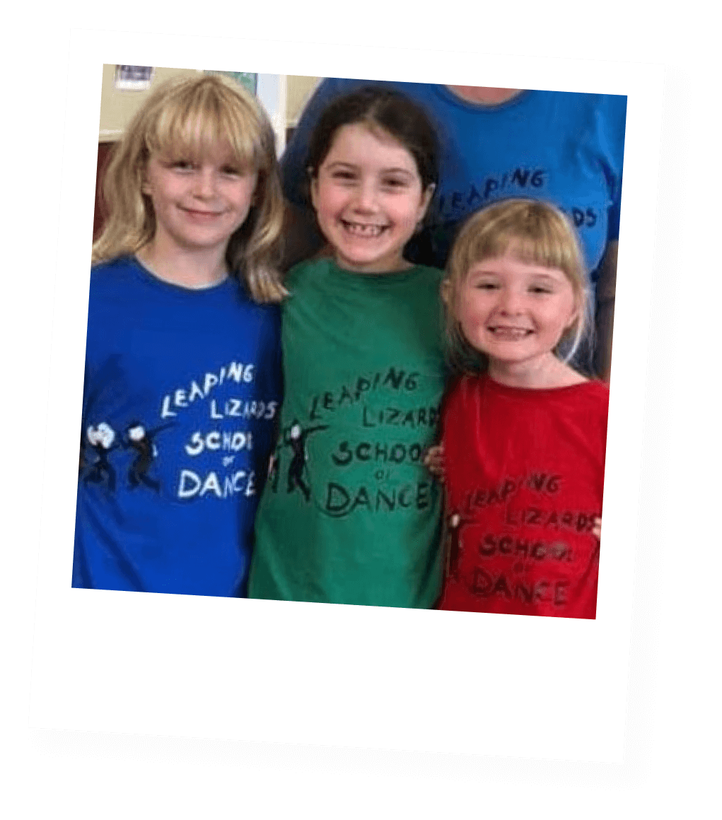 Three young girls smiling, wearing colorful shirts with the text "Leaping Lizards School of Dance" and a lizard graphic on each shirt, standing close together in a dance studio.