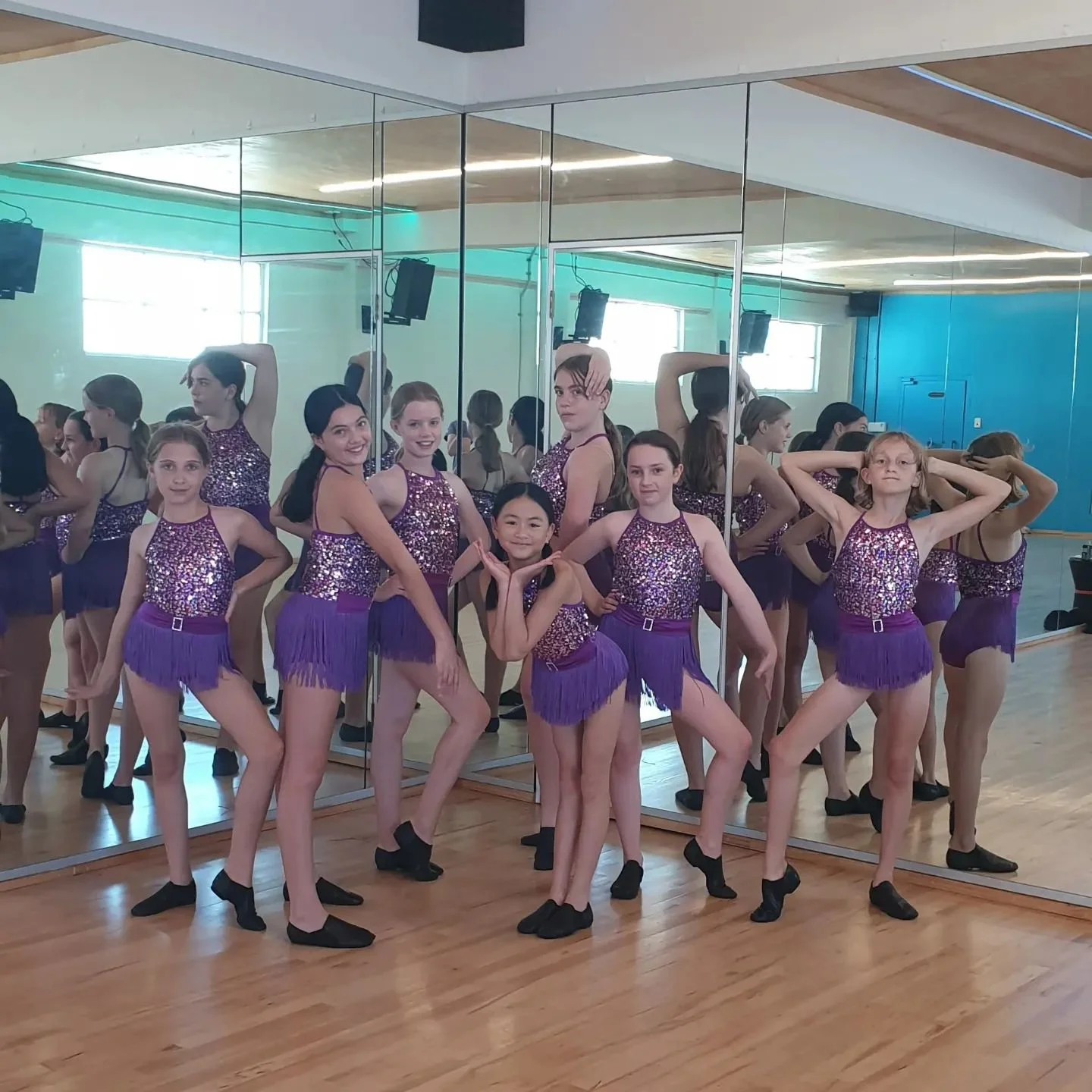 Group of young girls in purple sequined dance costumes standing in front of a mirror in a dance studio.