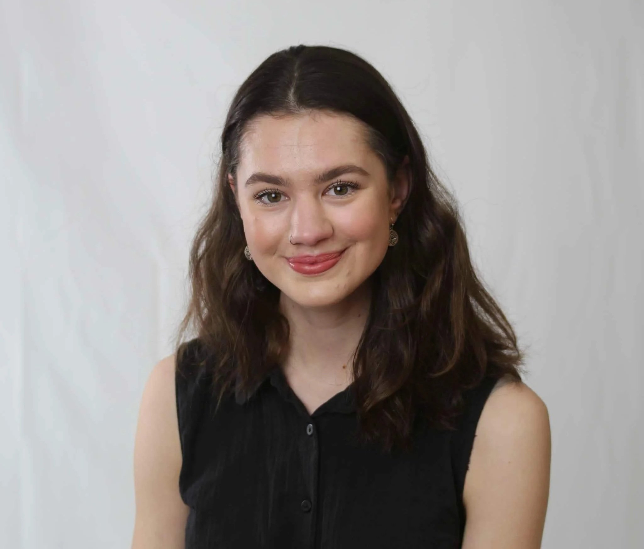 A young woman with brown wavy hair, wearing earrings and a black sleeveless top, smiling at camera against a plain white background.