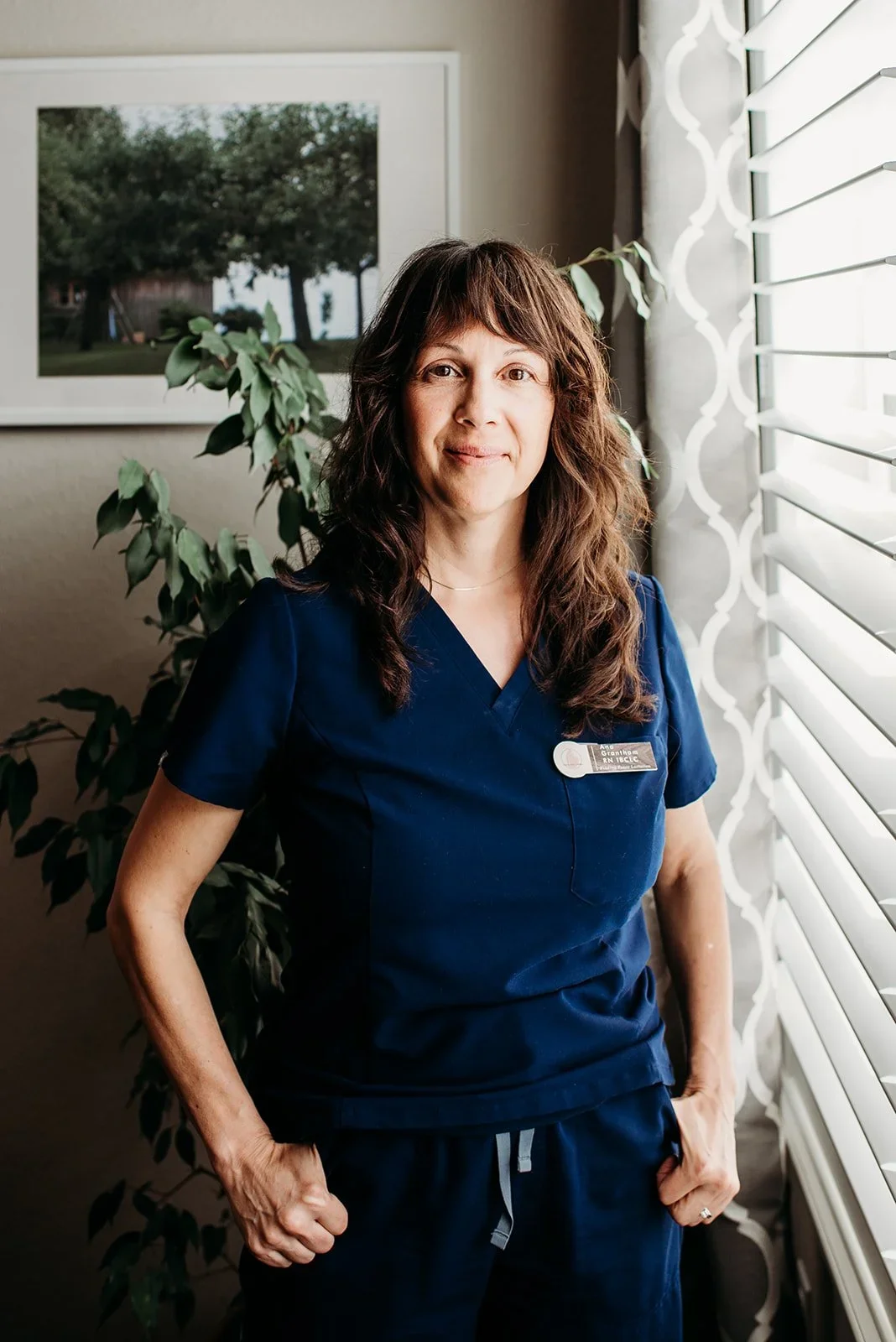 A woman in navy scrubs standing by a window with white blinds and a patterned curtain, with a framed landscape photograph on the wall behind her and a large green plant to her left.