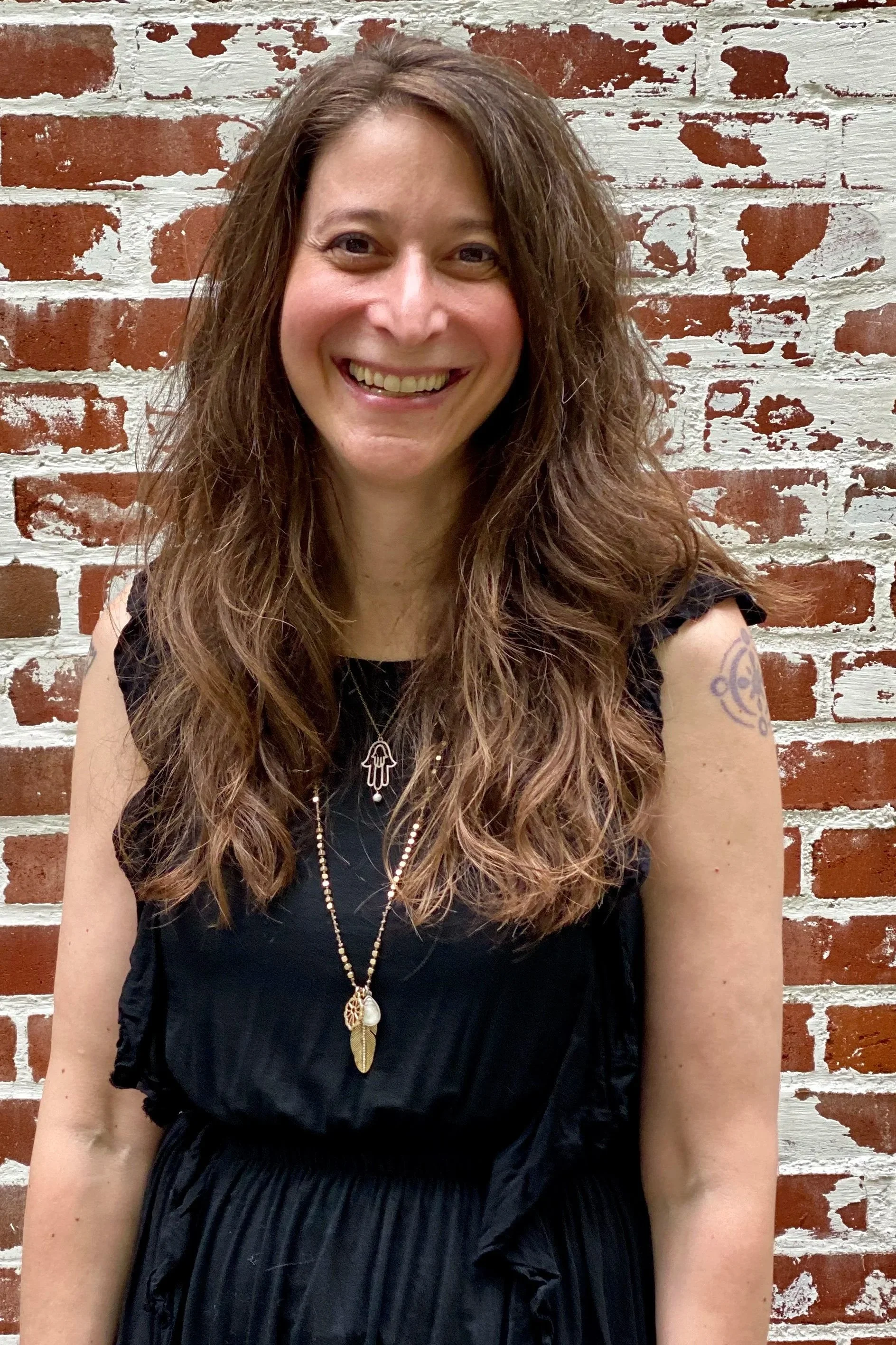 A woman with long, wavy hair smiling in front of a brick wall painted white.