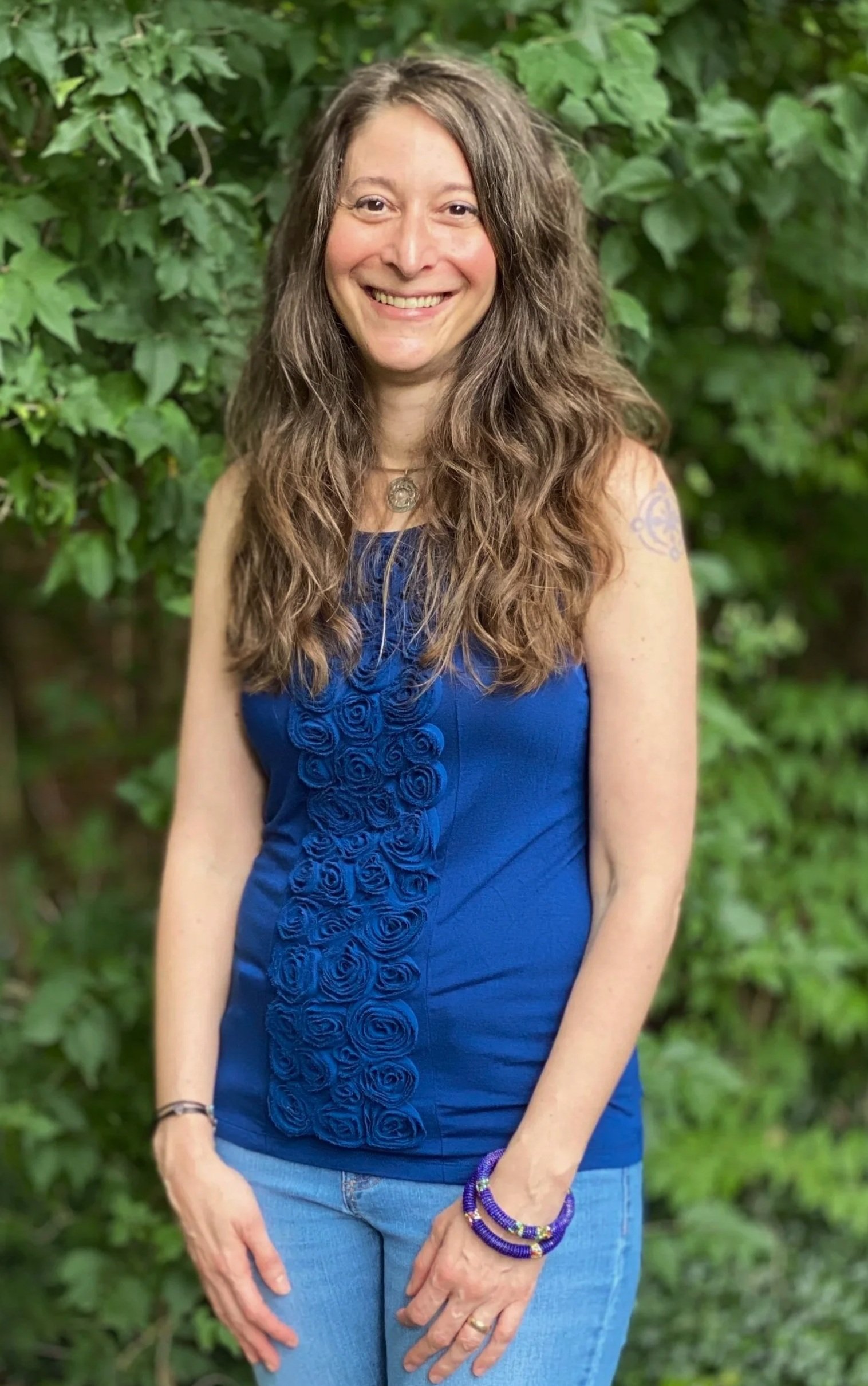 A woman standing outdoors in front of green foliage, smiling at the camera, wearing a blue sleeveless dress with a rosette pattern, blue jeans, and purple bracelets.