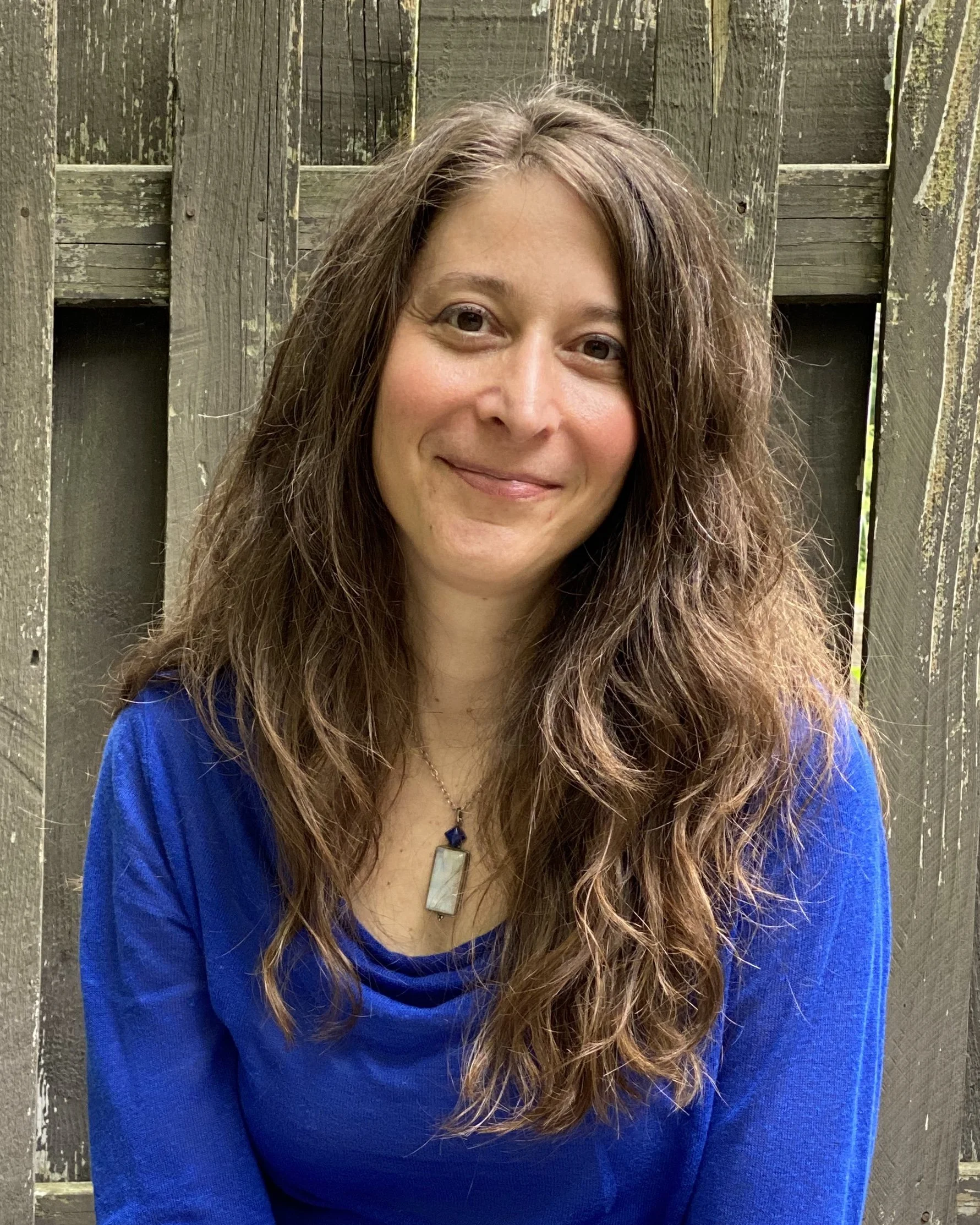 A woman with long, curly brown hair smiling in front of a weathered wooden fence, wearing a blue top and a rectangular pendant necklace.