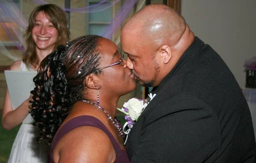 A couple is kissing at a wedding, with a woman in the background smiling and holding a book.