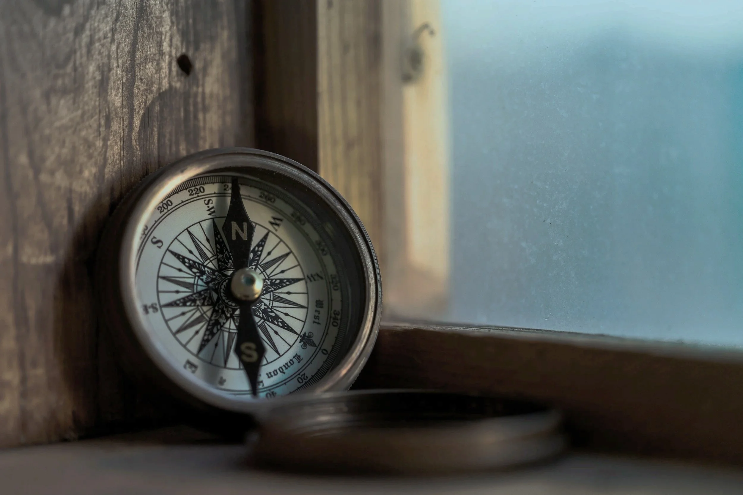 A compass lying on a wooden surface near a window with a blurred outdoor scene in the background.