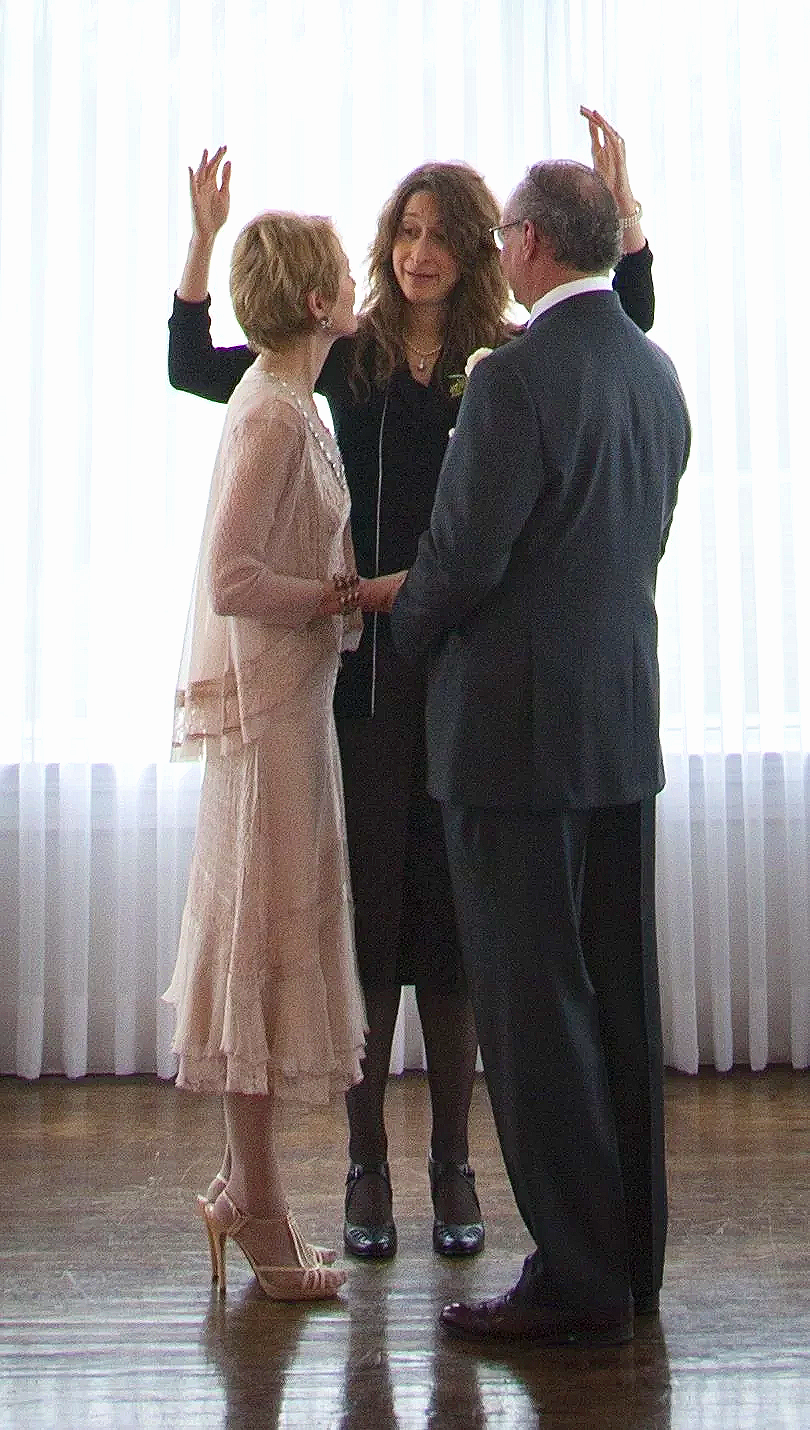 A woman and a man holding hands, facing each other, while Valerie officiates their wedding. The woman is wearing a pink dress and high heels, and the man is in a dark suit. Valerie, dressed in black, is standing between them with arms raised.
