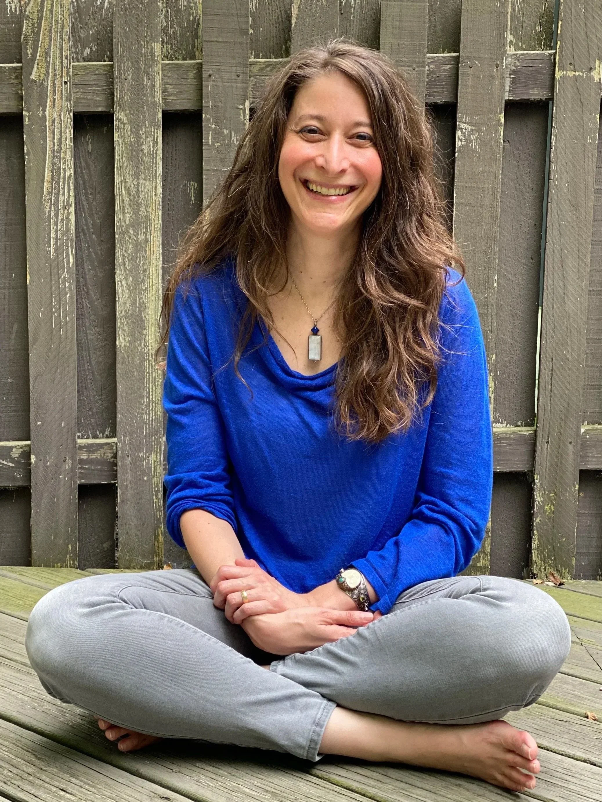 A woman with long, wavy brown hair sitting cross-legged on a wooden deck, smiling, against a wooden fence background, wearing a blue long-sleeve shirt, gray pants, and jewelry.