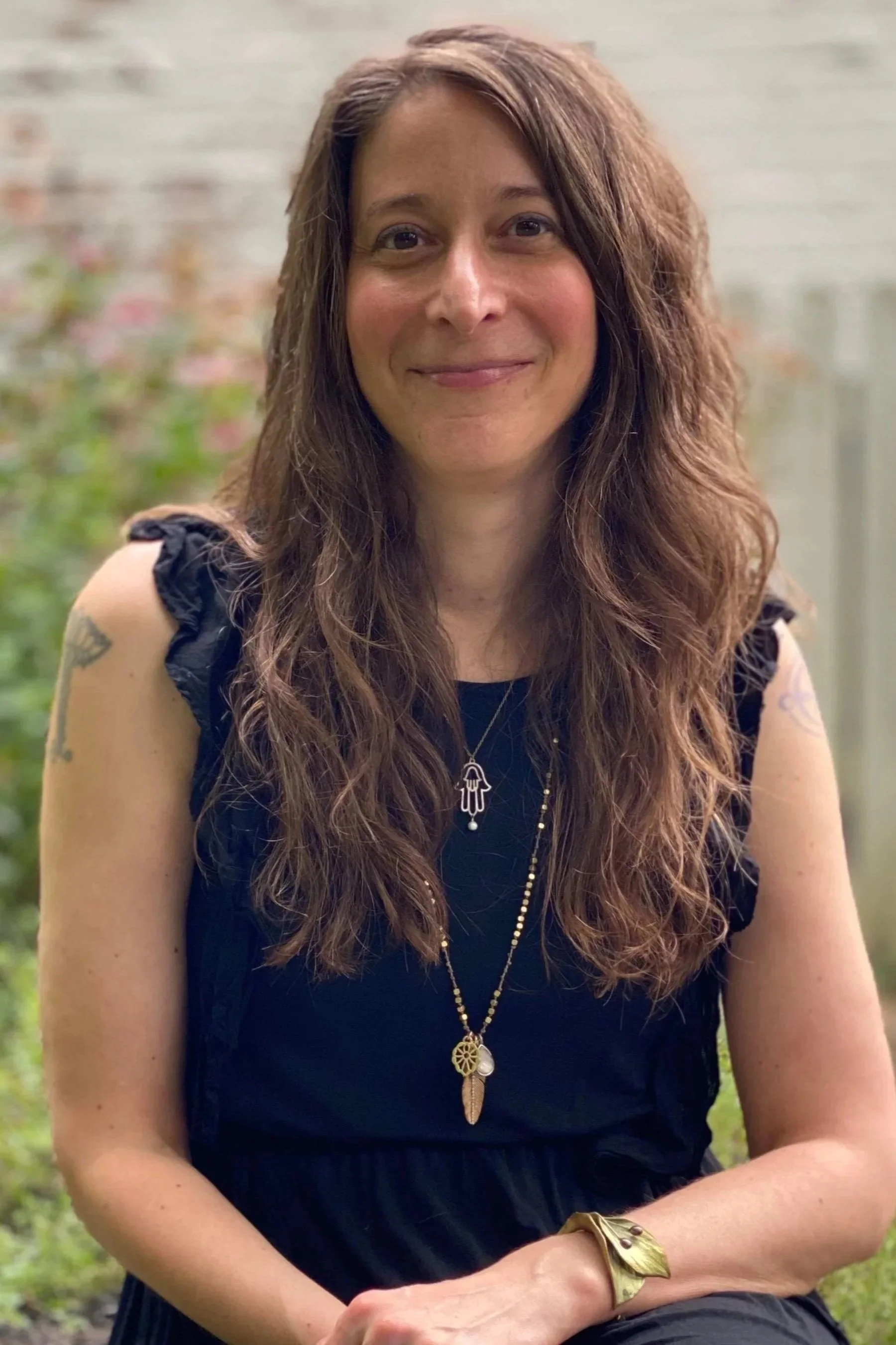 A woman with long wavy brown hair, smiling, wearing a black top with ruffled sleeves, a gold necklace with a pendant, a gold leaf bracelet, and tattoos on her arms, sitting outdoors with a blurred background of a brick wall and greenery.