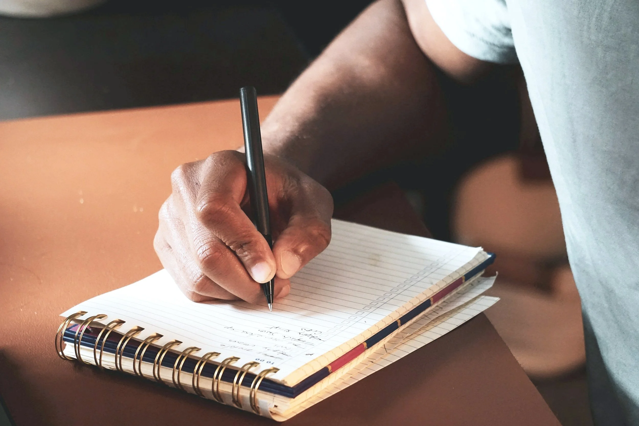 A person writing in a spiral notebook with a black pen, sitting at a desk.