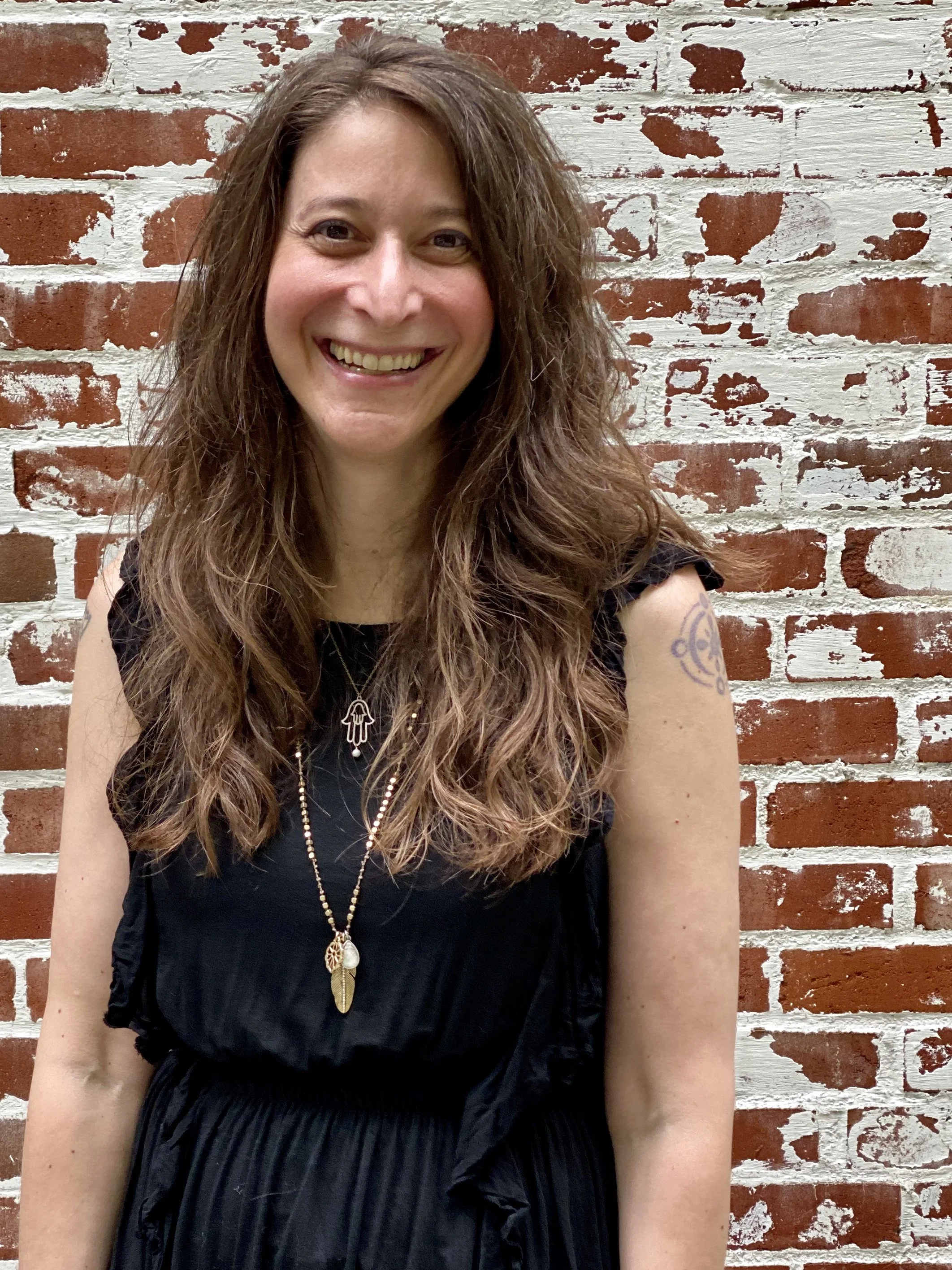 Smiling woman with long curly brown hair wearing a black sleeveless top, standing against a red brick wall with white paint accents.