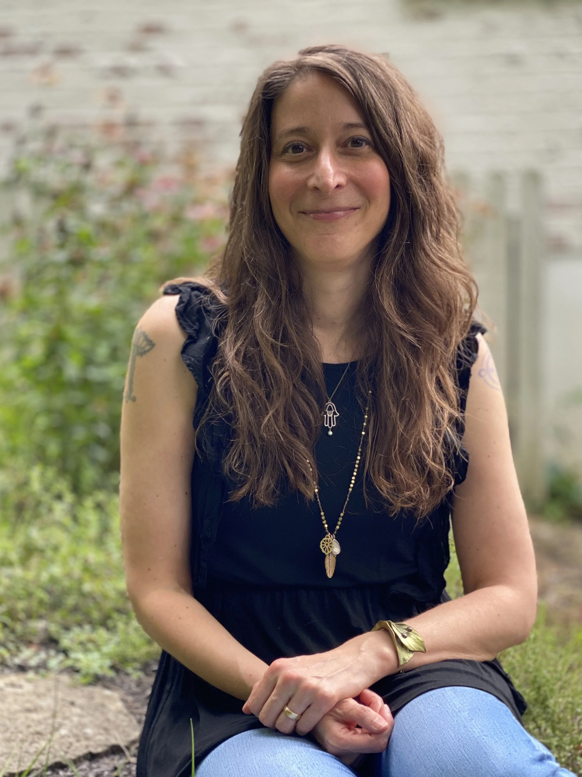 A woman with long wavy brown hair sitting outdoors in front of a blurred brick wall and greenery, wearing a black sleeveless top, blue jeans, and various jewelry, smiling at the camera.