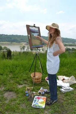 A woman wearing a wide-brimmed hat and white shirt standing outdoors painting on an easel by a river, with painting supplies on the ground.