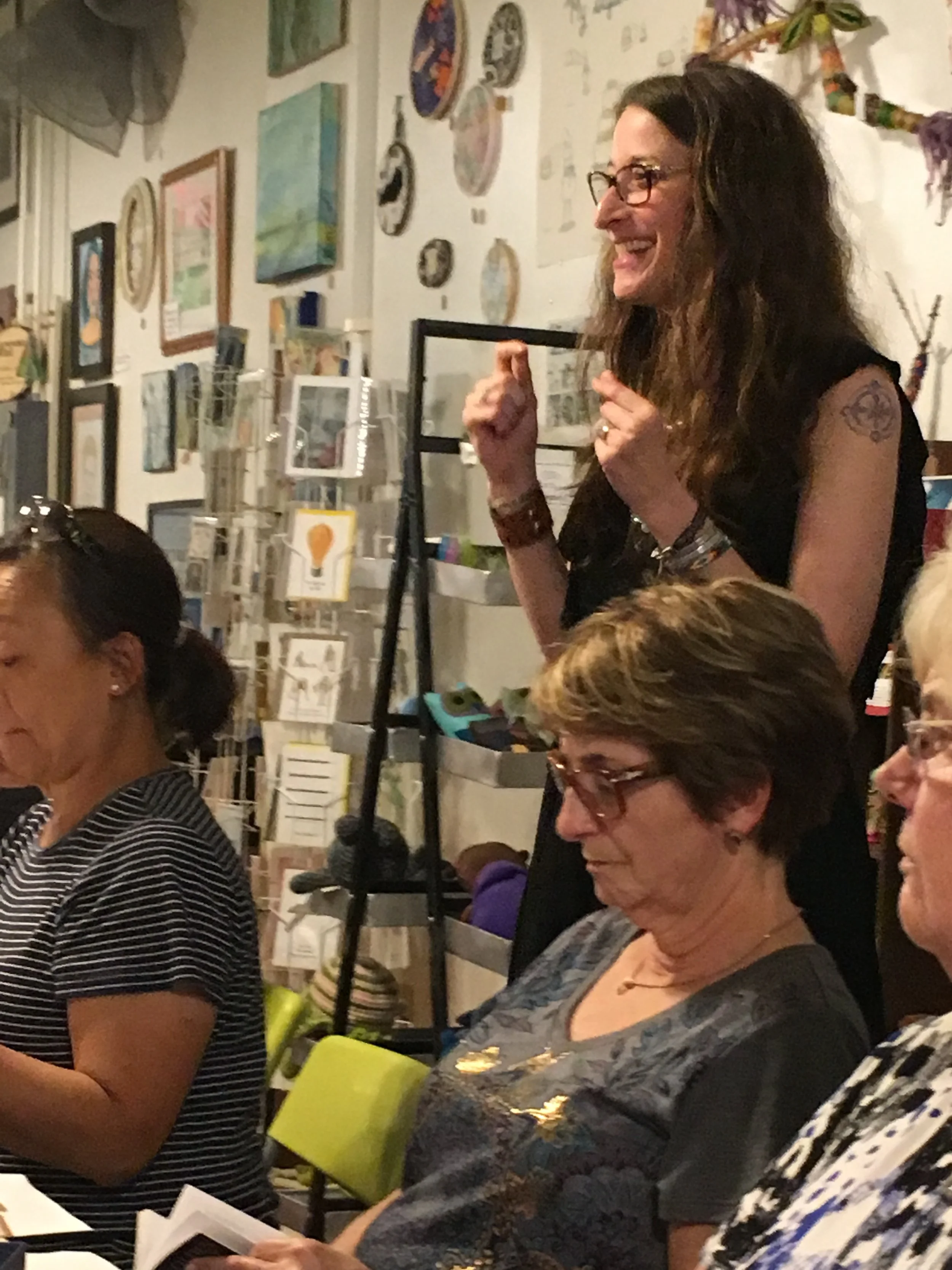 Smiling woman with glasses and long hair, standing and talking at a social gathering with people seated and reading, in an art-filled room.