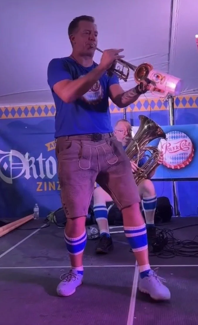 A man wearing a blue t-shirt and traditional Bavarian Lederhosen playing a trumpet on stage, with a band behind him. The stage has a banner with German text, and a person in the background is playing a tuba, wearing German socks and shoes.