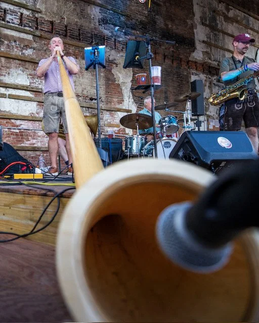Musicians performing on a stage with exposed brick wall, including a person playing a didgeridoo, a drummer, and a saxophonist; a microphone and sound equipment in the foreground.