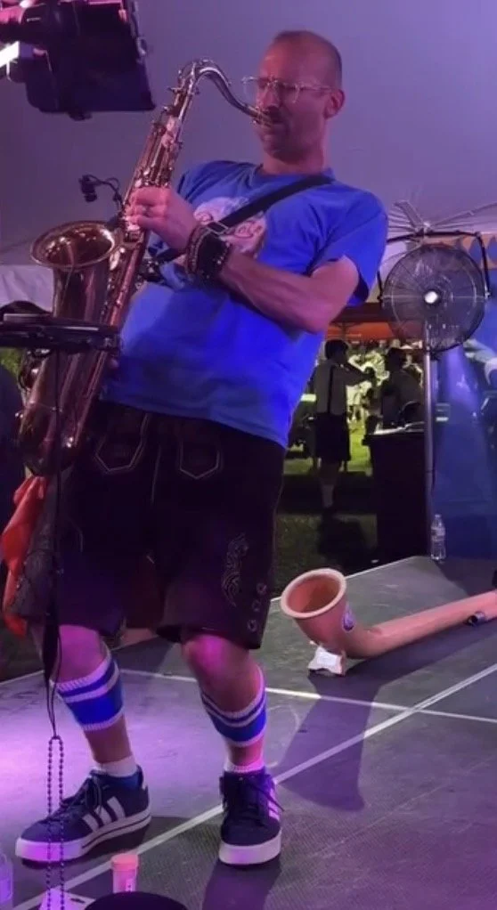 A man in a blue t-shirt, glasses, lederhosen and German socks is playing a saxophone on a stage, with an alphorn and a fan in the background.