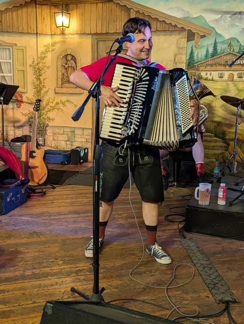 A man smiling and playing an accordion on stage with musical equipment and a scenic mural background.