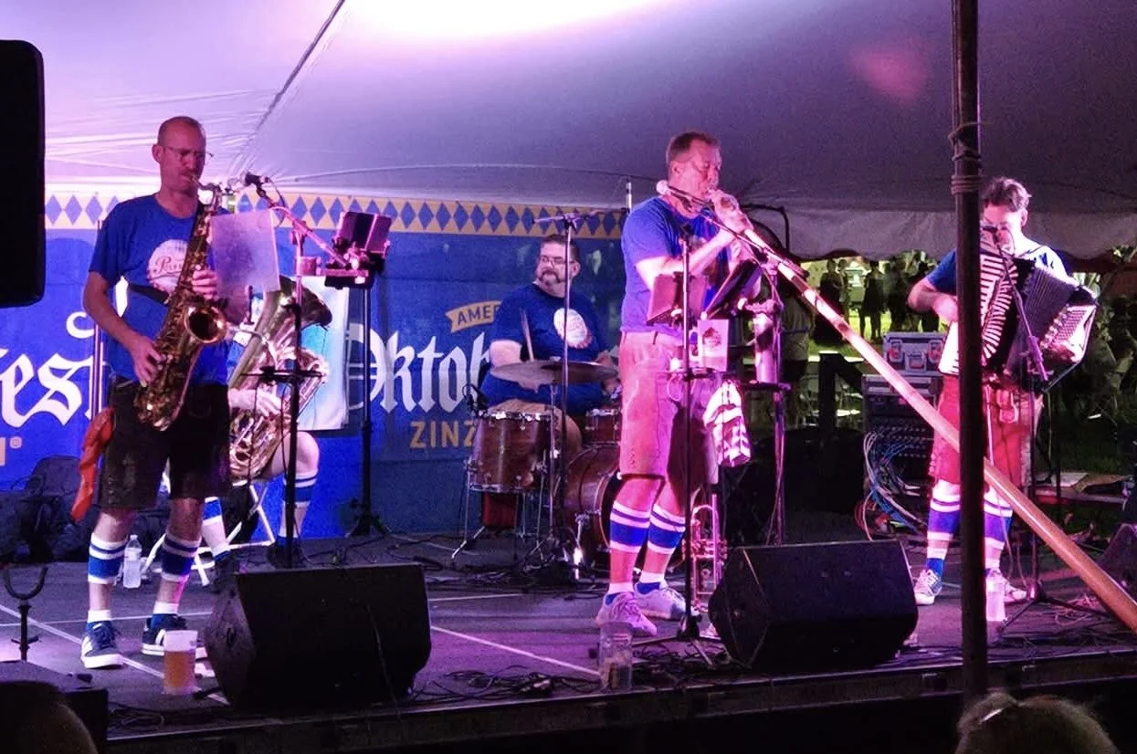 A band performing on stage at a festival inside a large tent, with members playing saxophone, drums, aphorn, and accordion, wearing traditional Bavarian style clothing including blue shirts, lederhosen, and German socks.