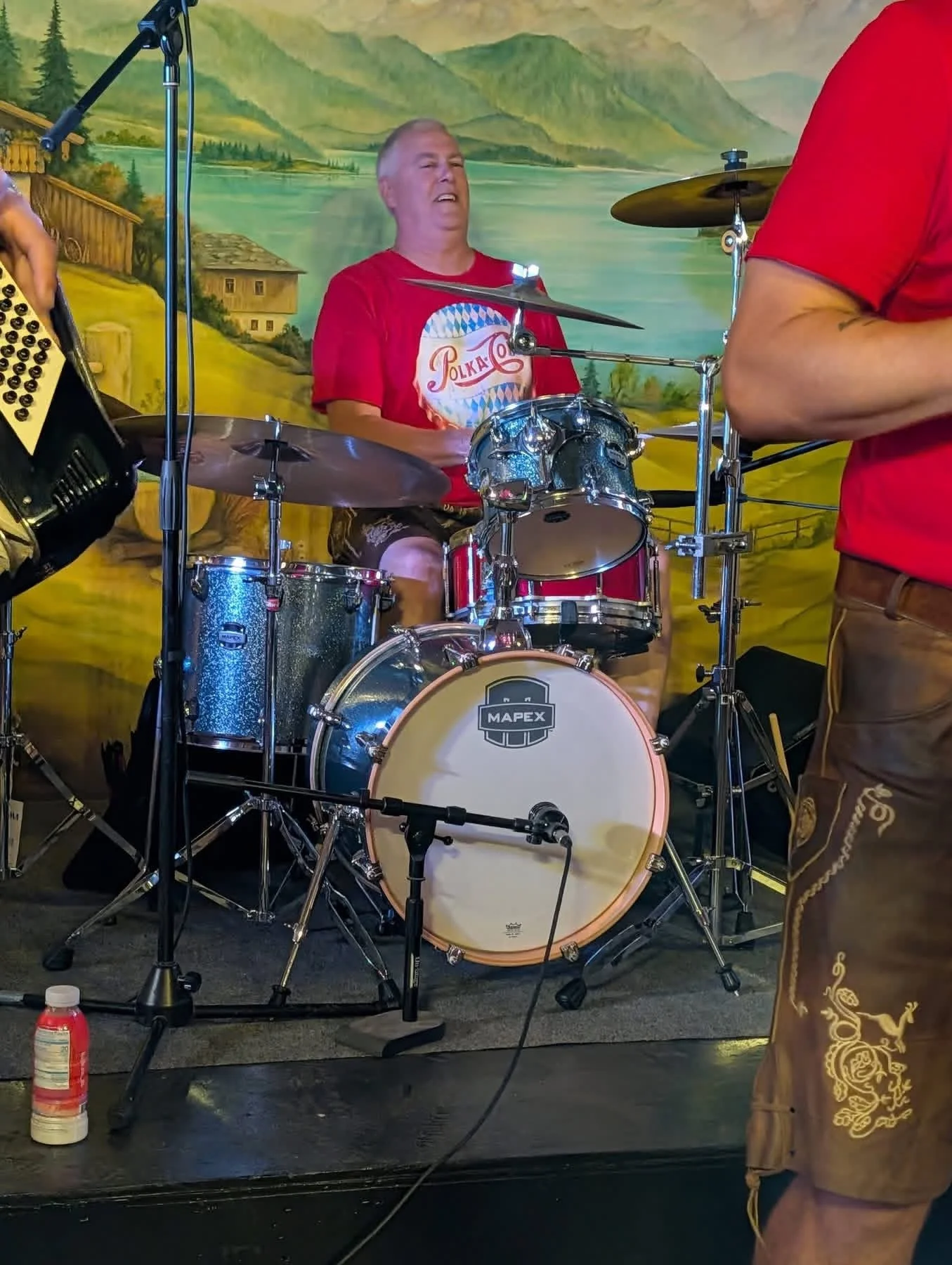 A man with short gray hair playing drums in a band, wearing a red t-shirt with the word 'Polka Cola' and a logo on it. He is sitting behind a drum set with a painted scenic mural of mountains, a lake, and houses in the background.