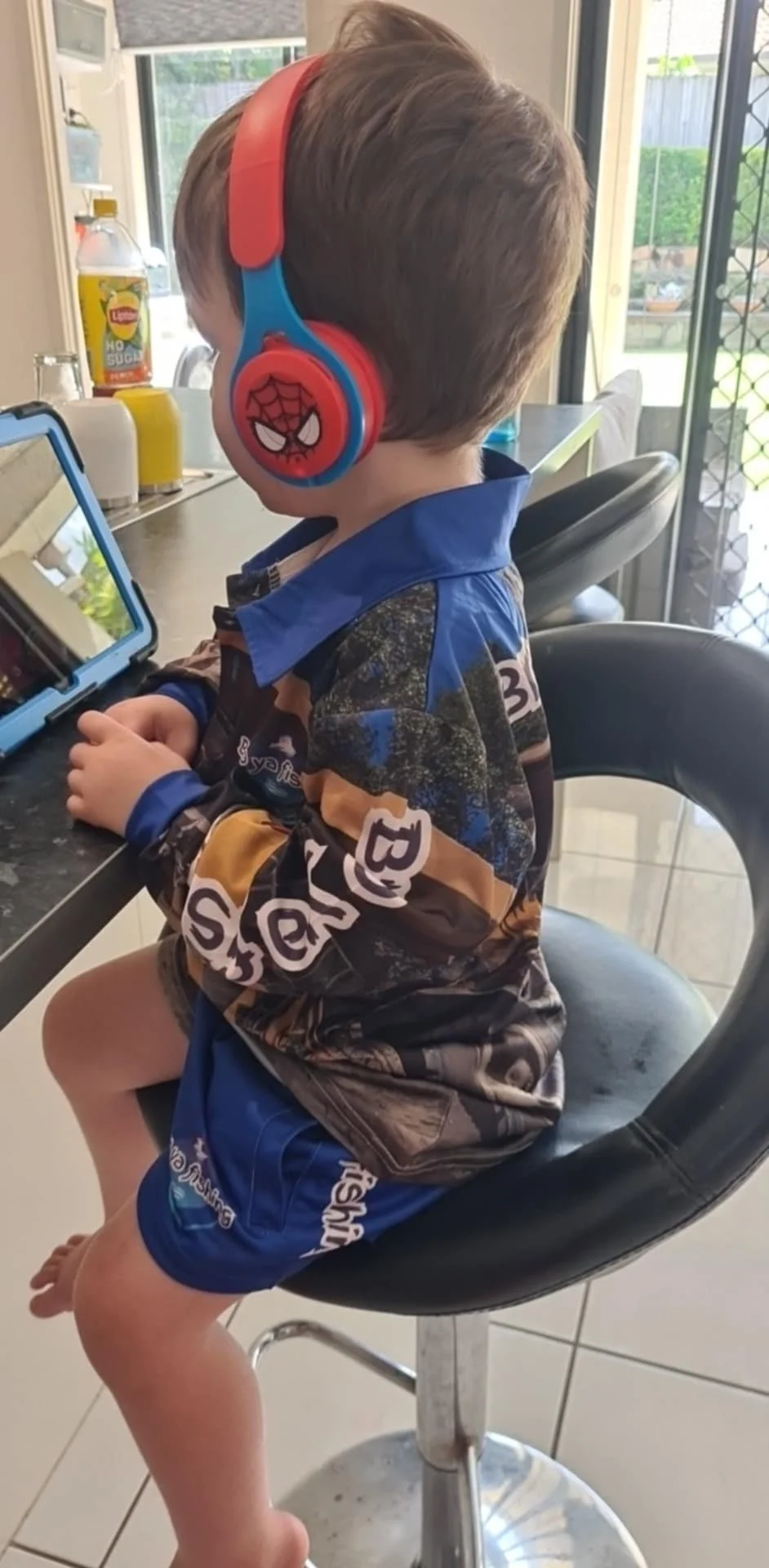 A young boy sitting on a black barstool, wearing a Spider-Man themed face mask on his cheek, headsets, a camouflage long-sleeve shirt, and blue shorts, looking at a tablet on a counter inside a house with a sliding door and outdoor yard visible in th