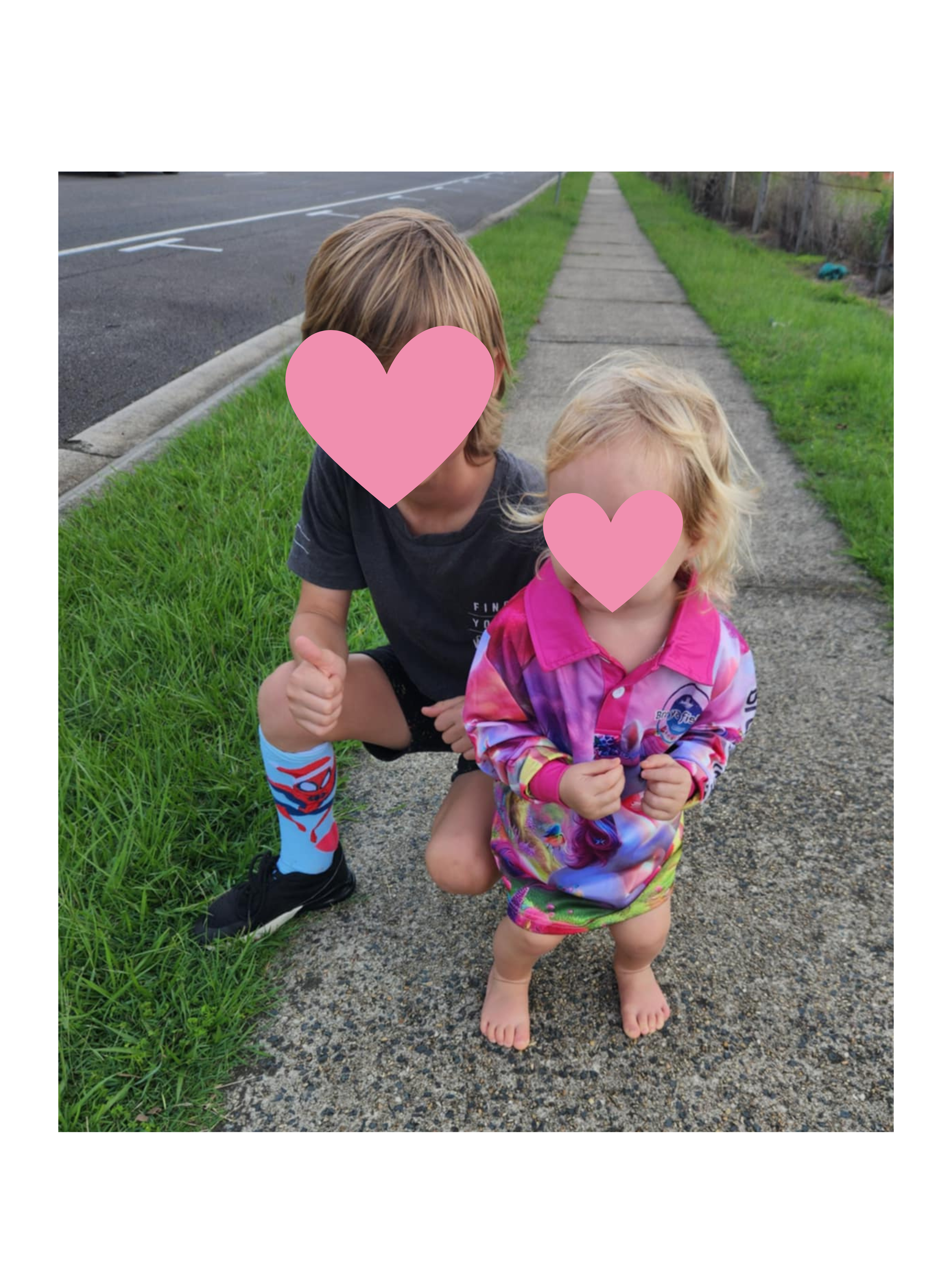 Two children standing on a sidewalk, with a grassy area on one side and a street on the other, posing for the photo. Their faces are covered with pink heart overlays.