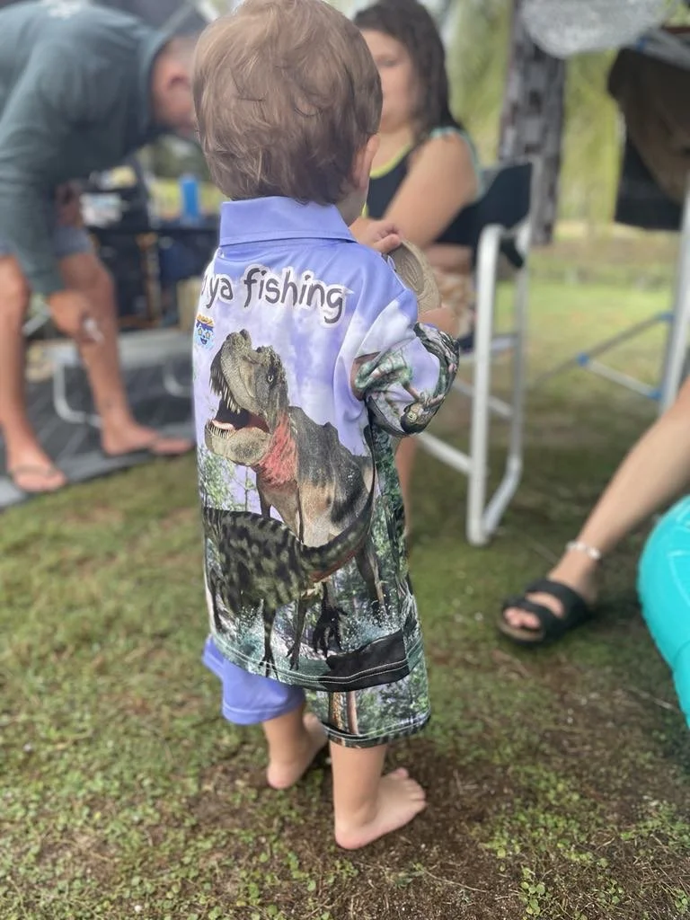 Child in a dinosaur-themed shirt standing barefoot on grass at an outdoor gathering, holding a goody bag, with adults seated nearby.