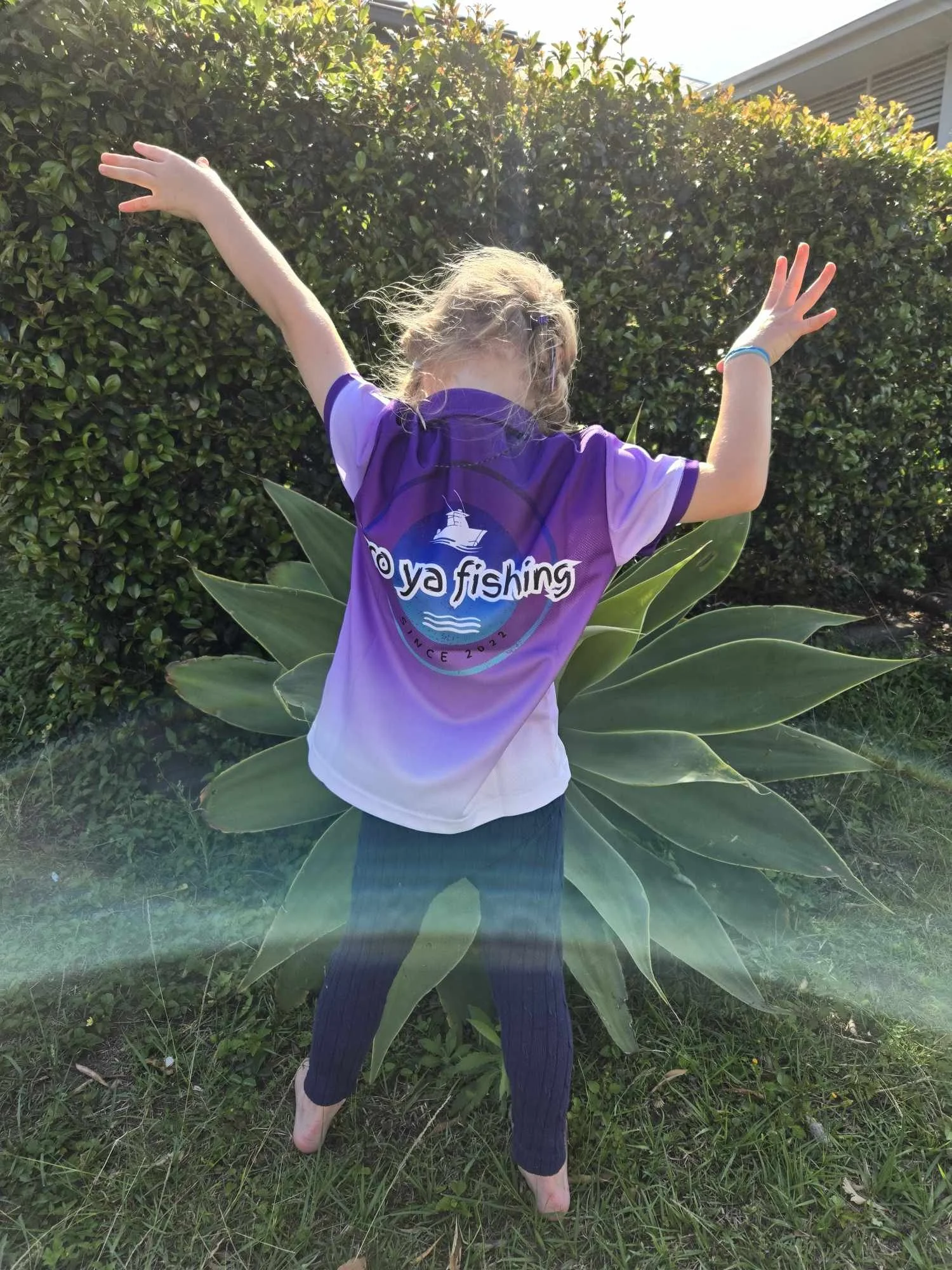 Child standing outdoors with back to camera, arms raised, wearing a purple and white shirt with 'Oya Fishing' logo, in front of a large leafy plant and green hedge.
